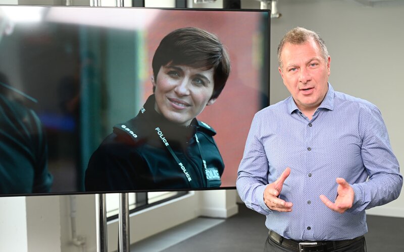 Jed Mercurio stands next to a tv playing Line of Duty